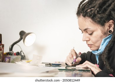 Beautiful Young White Girl With Dreadlocks Hispanic Latina Art Director In Her Art Studio, Cutting Cardboard On A Table, Illuminated By A Lamp And With Working Implements Around Her.