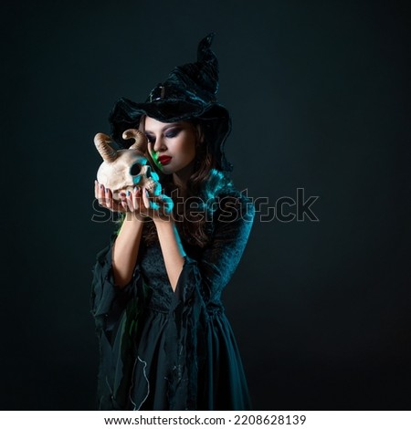 A beautiful young witch holds a horned demon skull near her face and looks at you with a sly look, an invitation to a Halloween party, a witch costume, a black dress and a hat.