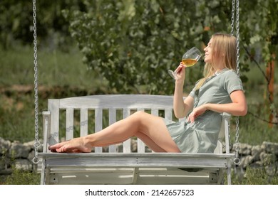 Beautiful Young Woman Against Summer Green Park. Wine Handmade From White Grape. Young Woman Tasting Wine Or Grapes Juice. Hipster Girl Sitting On A Wooden Swing.