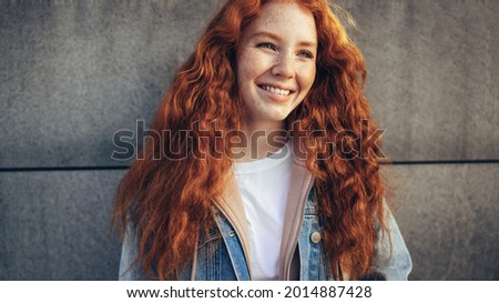 Beautiful young woman against wall. Student with red dyed hair looking away and smiling
