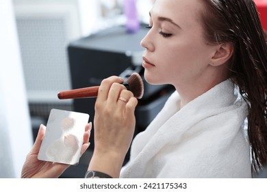 A beautiful young woman applies blush to her face with a makeup brush. Professional makeup in a beauty salon. Form. Cosmetics. The foundation for perfect makeup. - Powered by Shutterstock