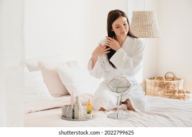 Beautiful Young Woman Applying Beauty Oil On Her Long Hair For Care And Treatment. Girl Relaxing On Her Bed In Spa Bathrobe, Preparing For Sleep. Hair Growth Volume Effect