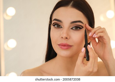 Beautiful young woman applying black eyeliner indoors. Space for text