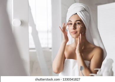 Beautiful Young Woman Applying Cream On Her Face In Bathroom, Reflecting In Mirror. Anti Age Eye Cream Concept