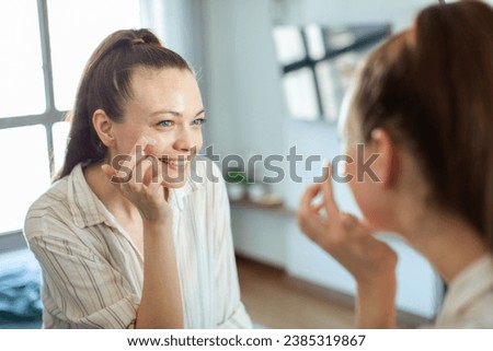Beautiful young woman applying facial cream for healthy skin with mirror