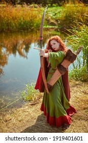 Beautiful young woman archer with magnificent long red hair in a historical celtic dress is aiming a bow near the lake. Historical reconstruction. 