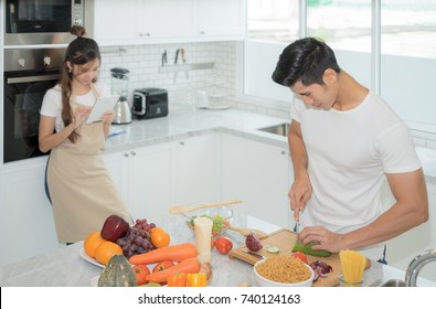 Beautiful Young Woman Asian Is Using A Digital Tablet And Smiling While Her Man Is Cooking In Kitchen At Home