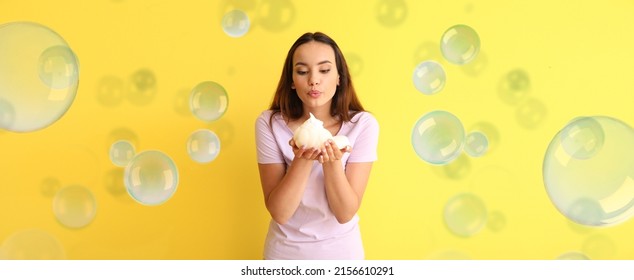 Beautiful young woman with bast wisp and soap bubbles on yellow background