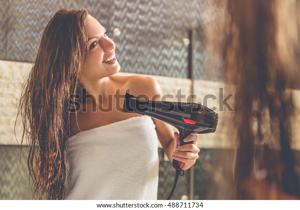 Beautiful young woman\
in bath towel is using a hair dryer and smiling while looking into\
the mirror in\
bathroom