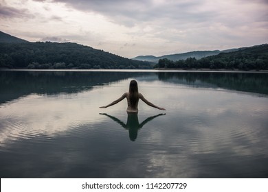 Beautiful Young Woman Bathes In A Lake. It Stands At The Bottom Of The Waist In The Water. Her Hands Are Raised Above The Water. A Silhouette Of Her Body, Her Hair Is Loose. Reflection In Water.