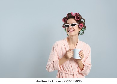 Beautiful young woman in bathrobe with hair curlers holding cup of drink on light grey background. Space for text - Powered by Shutterstock