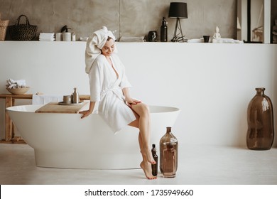 Beautiful Young Woman In Bathrobe Sitting On The Edge Of Bathtub. Self Care Concept.