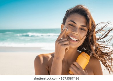 Beautiful Young Woman At Beach Applying Sunscreen On Face And Looking At Camera. Beauty Latin Girl Applying Suntan Lotion At Sea. Portrait Of Happy Woman With Healthy Skin Applying Sunblock On Cheek.