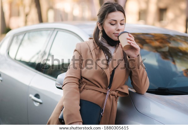 Beautiful young woman in black face\
mask stand by the car and drink coffee. Happy young woman have a\
break. Female pulled off mask for drinking cappucino\
outdoors