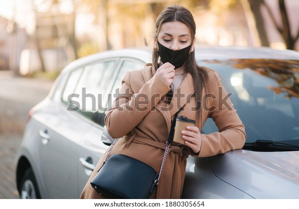 Beautiful young woman in black face
mask stand by the car and drink coffee. Happy young woman have a
break. Female pulled off mask for drinking cappucino
outdoors