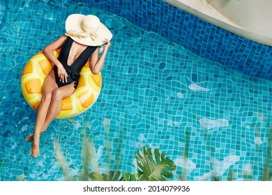 Beautiful Young Woman In Black Swimsuit Covering Face With Straw Hat When Lying On Floating Ring In Swimming Pool