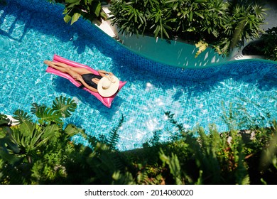 Beautiful Young Woman In Black Swimsuit And Straw Hat Floating On Pink Mattress In Swimming Pool