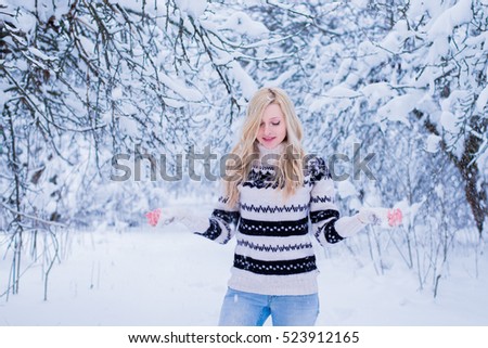 Beautiful young woman with blond hair is dressed in a warm black and white sweater on the snowy background in the winter forest. / Beautiful young woman throwing snow and smiling. Christmas theme