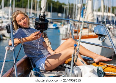Beautiful young woman blonde girl eyes closed, relaxing with a book, sitting on the deck of a boat yacht listening to music on earphones and a mobile cell phone in summer sunshine