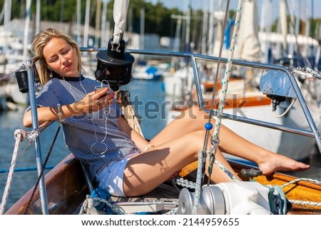 Beautiful young woman blonde girl eyes closed, relaxing with a book, sitting on the deck of a boat yacht listening to music on earphones and a mobile cell phone in summer sunshine