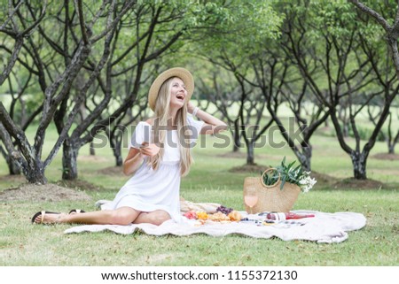 Beautiful young woman with blonde hair in straw hat and white dress sitting on the plaid in the park. It's summer picnic