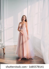 Beautiful Young Woman. Blonde Long Flowing Hair. Elegant Airy Peach Beige Ball Gown. Evening Long Pink Trendy Tulle Dress. Backdrop White Window Vintage Classic Room. Prom Holiday Image. Stylish Look