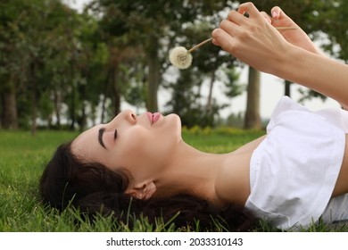 Beautiful young woman blowing dandelion while lying on green grass in park. Allergy free concept