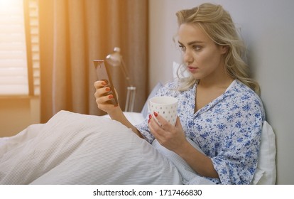 Beautiful Young Woman Browsing Smartphone And Holding Mug Of Hot Beverage Sitting On Bed Under Warm Blanket.