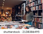 Beautiful young woman buying books at a bookstore and reading one. Geeky woman reading a book with a bookshelves in background.