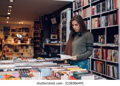 Beautiful young woman buying books at a bookstore and reading one. Geeky woman reading a book with a bookshelves in background.