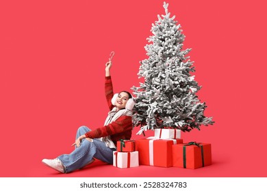 Beautiful young woman with candy canes and gift boxes near Christmas tree on red background - Powered by Shutterstock