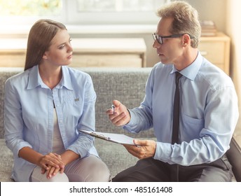 Beautiful Young Woman In Casual Clothes Is Sitting On The Couch At The Psychotherapist. Handsome Middle Aged Doctor Is Holding A Folder And Talking To Her