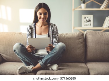 Beautiful Young Woman In Casual Clothes Is Using A Digital Tablet And Smiling While Sitting On Couch At Home