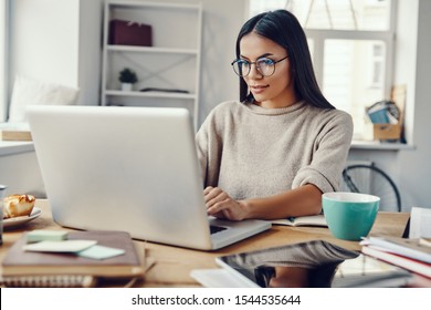 Mulher jovem e bonita em roupas casuais usando laptop e sorrindo enquanto trabalhava dentro de casa