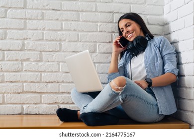 Beautiful young woman in casual wear is using a laptop and smiling, sitting against white brick wall