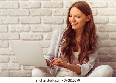 Beautiful young woman in casual wear is using a laptop and smiling, sitting against white brick wall