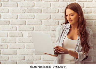 Beautiful young woman in casual wear is using a laptop and smiling, standing against white brick wall