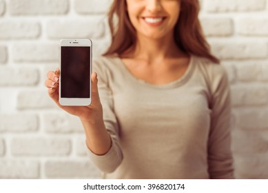 Beautiful young woman in casual wear is presenting a smartphone, looking at camera and smiling, standing against white brick wall, close up