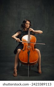 Beautiful young woman, cellist sitting on chair with cello, looking at camera on vintage green background. Cultural events with classic music performances. Concept of classical art, retro style, music