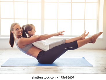 Beautiful young woman and charming little girl are smiling while doing yoga together in fitness hall
