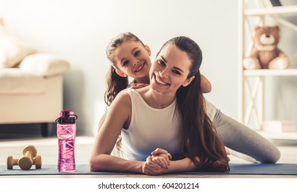 Beautiful young woman and charming little daughter are looking at camera and smiling while working out at home