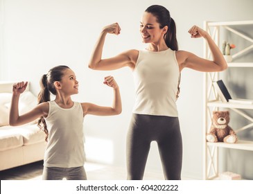 Beautiful young woman and charming little daughter are showing their biceps and smiling while working out at home