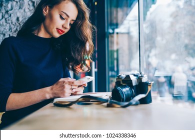 Beautiful young woman chatting online with colleague on modern mobile phone sitting at table with vintage camera in coffee shop interior in wifi zone.Copy space area for your advertising text message