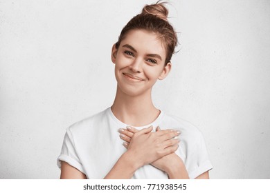 Beautiful Young Woman With Cheerful Friendly Expression, Keeps Hands On Heart, Demonstrates Her Love And Sympathy To Peope Who Surround Her, Glad To Recieve Words Of Encouragment, Isolated On White