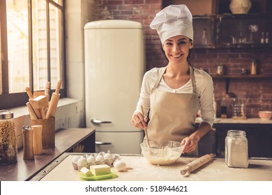 Beautiful Young Woman In Chef Hat Is Mixing Batter, Looking At Camera And Smiling While Baking In Kitchen At Home