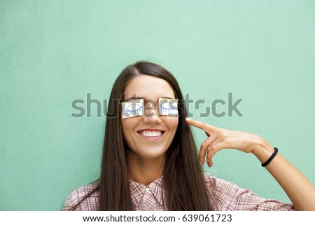 Beautiful young woman with chocolate long hair smiling happily with paper stickers with painted eyes on closed sleeping eyes, finger point at sticker on the eyes.