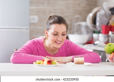 Beautiful young woman choosing between fruits and sweets in her kitchen.