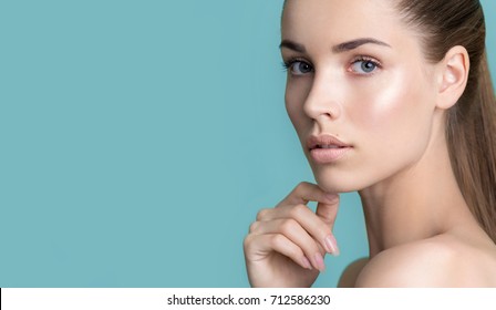 Beautiful young woman with clean perfect skin. Portrait of beauty model with natural nude make up and touching her face. Spa, skincare and wellness. Close up, blue background, copyspace.