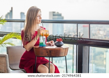 Beautiful young woman with cocktails rests at luxury rooftop restaurant. Elegant female lady in red dress with mocktails at sky bar terrace looking at modern city skyline. Skyscrapers on background.