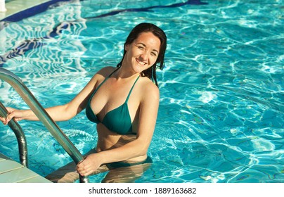 Beautiful young woman coming out of pool, holding stairs of swimming pool. Seductive model in blue swimsuit with dark hair posing at pool, looking at camera. Concept of pool outdoors.